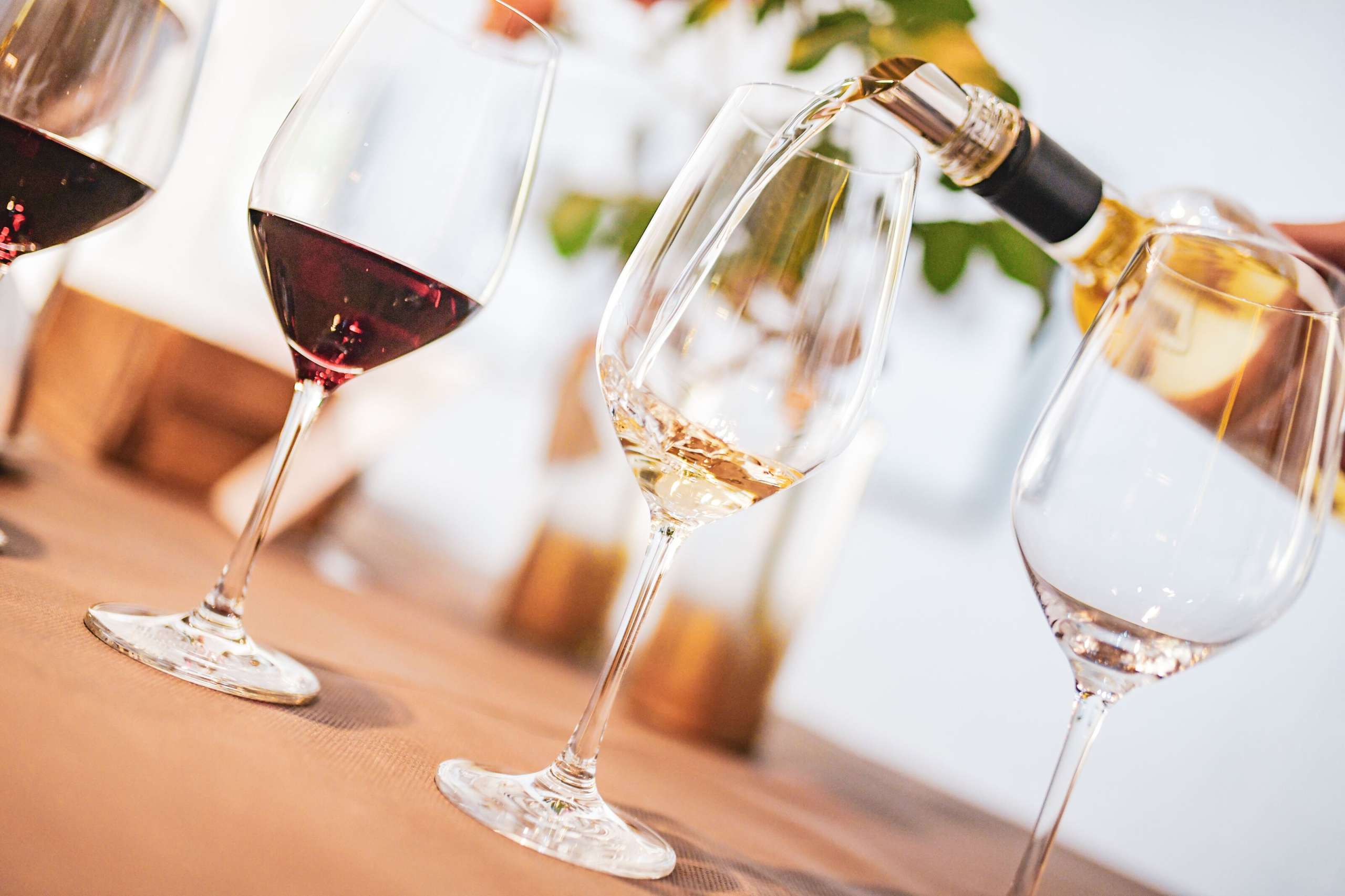 Es stehen vier Weingläser auf einem Tisch. Zwei davon sind mit Rotwein befüllt, eines ist Leer und in einem wird Weisswein eingeschenkt.