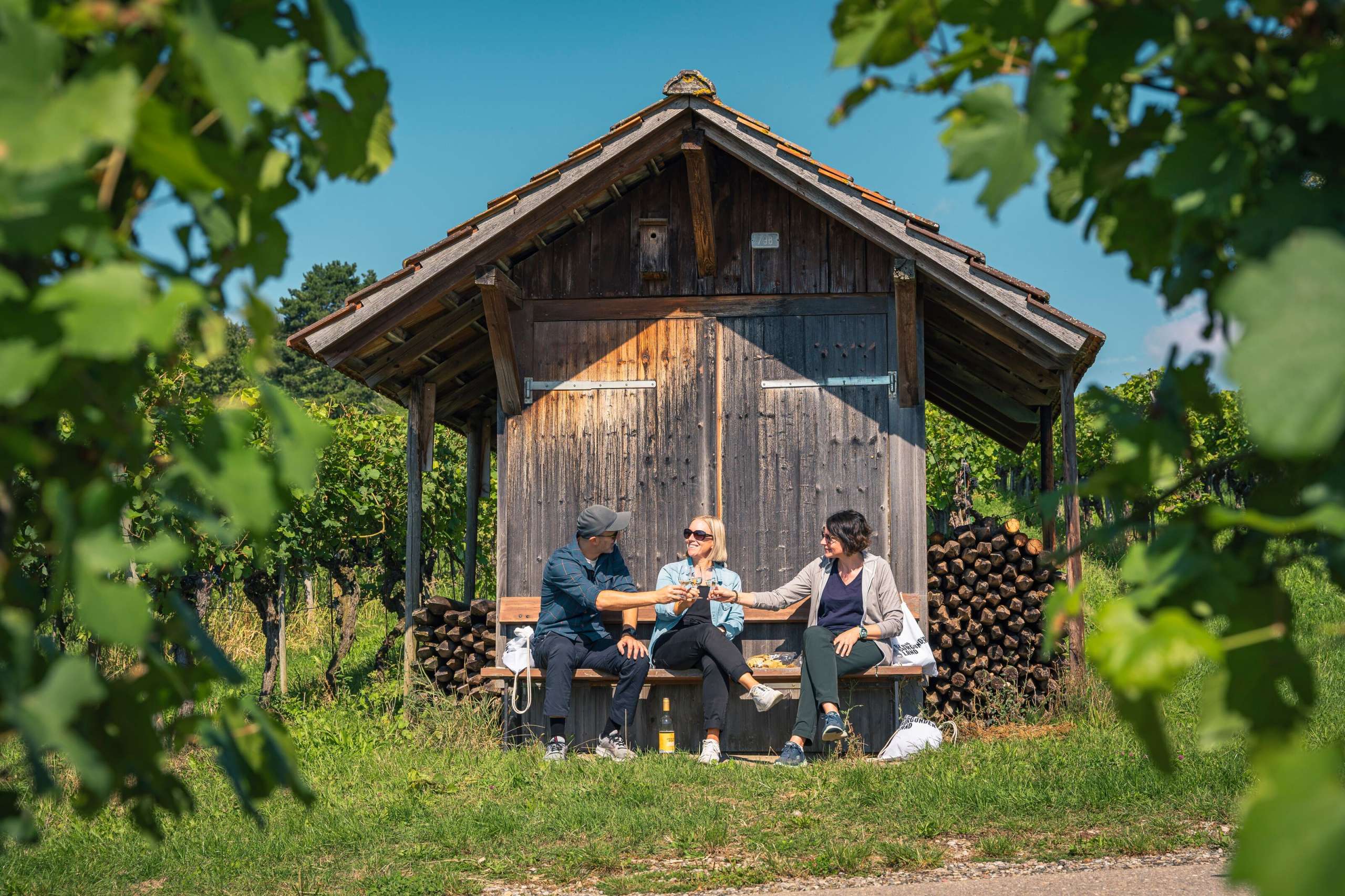 Drei Personen sitzen auf einer Holzbank vor einem Räbhüüsli in den Reben. Sie stossen mit einem Glas Weisswein an.