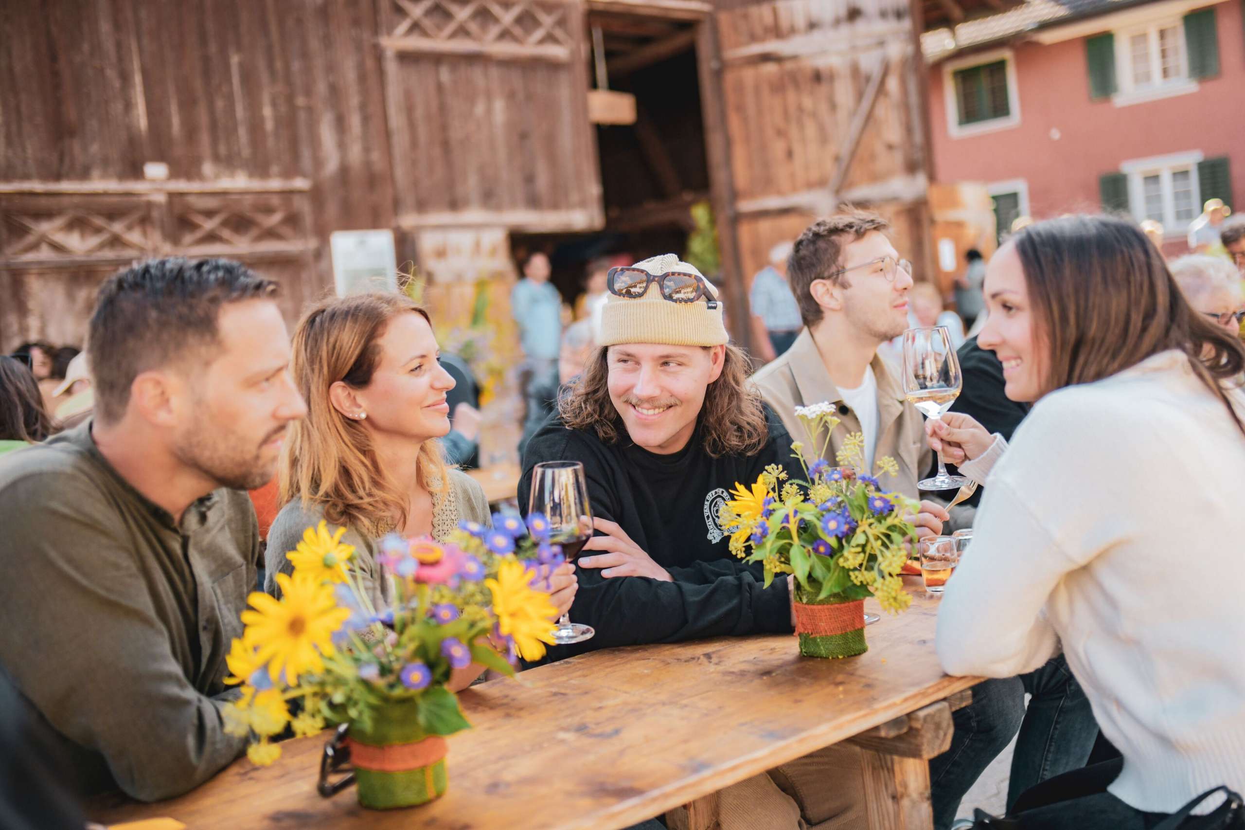 Eine Gruppe an Freunden sitzt an einem Festbank, welcher mit Blumen geschmückt ist. Im Hintergrund ist eine Scheune zu sehen. Die Freunde stossen mit einem Glas Wein an.