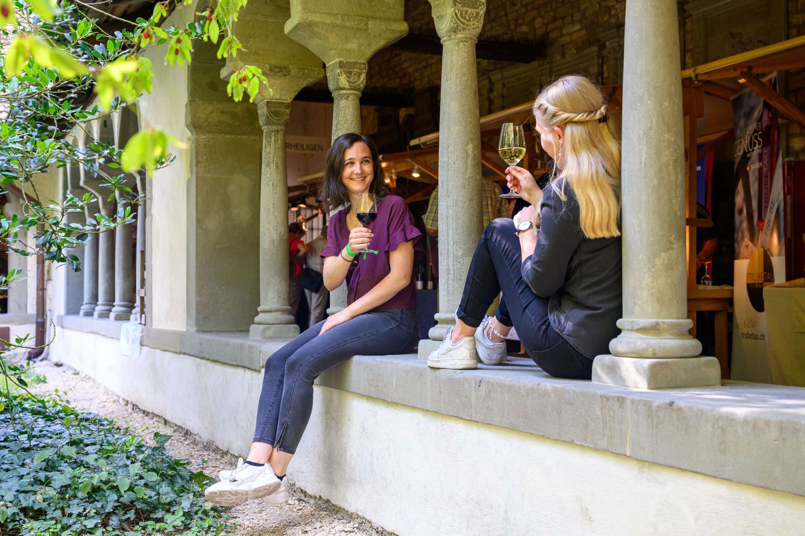 Zwei Freundinnen sitzen auf einer Steinmauer im Kreuzgang. Beide halten ein Glas Wein in der Hand.