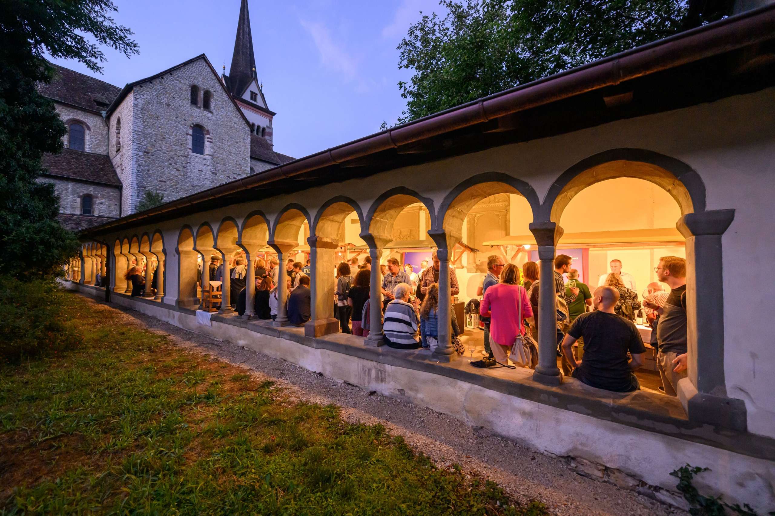 Diverse Stände, wo man Wein degustieren kann, sind im Kreuzgang zu Allerheiligen aufgestellt. Personen stehen an den Stände und trinken Wein.