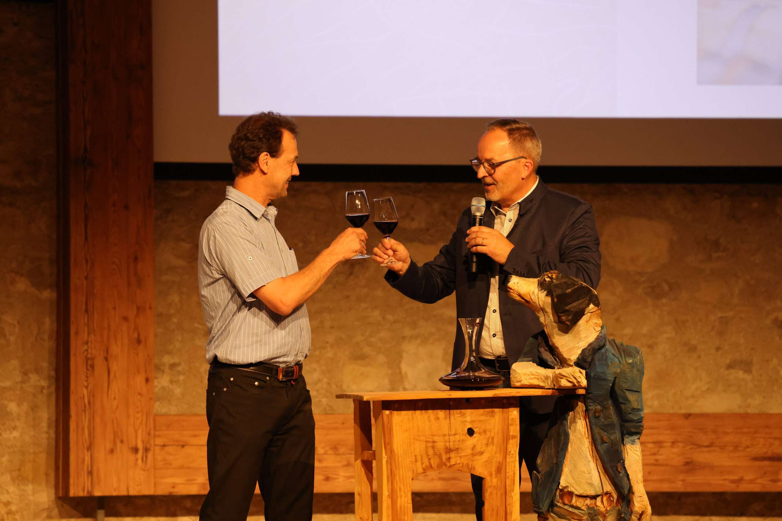 Zwei Männer stehen auf einer Bühne und stossen mit einem Glas Rotwein an.