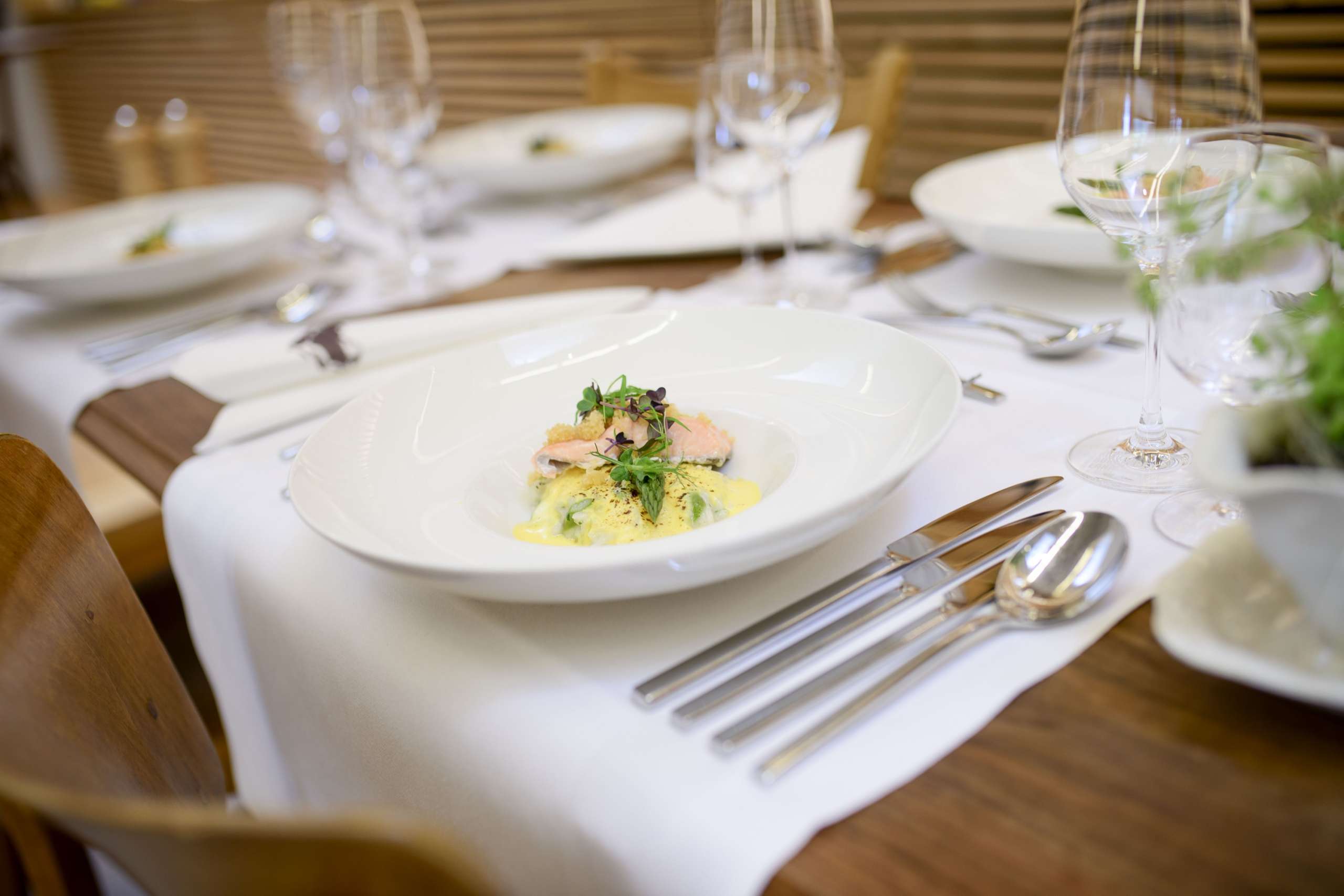 Ein Holztisch ist mit weissen Läufern gedeckt. Auf dem Tisch steht ein weisser Teller mit einem Fischgericht.